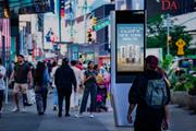 A busy New York City sidewalk with pedestrians walking past a LinkNYC digital kiosk displaying a Corona beer advertisement. The kiosk stands prominently in the foreground against a backdrop of bright city lights, storefronts, and traffic, illustrating digital out-of-home advertising in a high-footfall urban environment.
